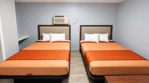 Habitación de hotel con dos camas, cada una con una colcha naranja y almohadas blancas, piso de madera y un equipo de aire acondicionado montado en la pared.