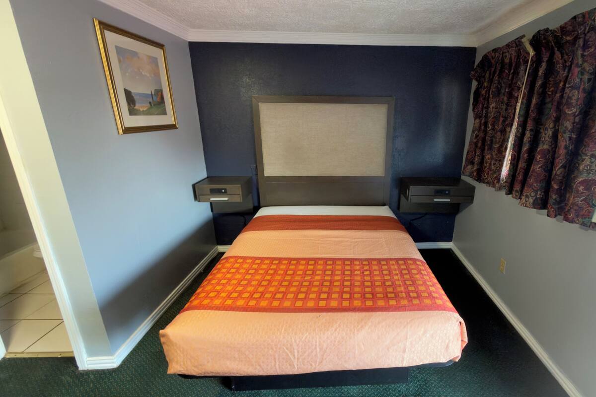 A hotel room with a bed, two bedside tables, a headboard, a window with curtains, and a framed picture on the wall.