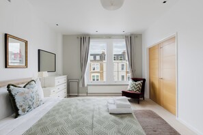Quarto com cama, cadeira, guarda-roupa, janela com cortinas e vista para edifícios.