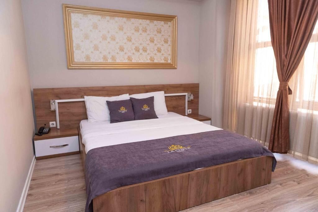 Hotellihuone, jossa on puinen sängynpääty, valkoisilla ja tummilla tyynyillä varustettu sänky, yöpöytä, jossa on puhelin, ja seinällä oleva kehystetty kuva.