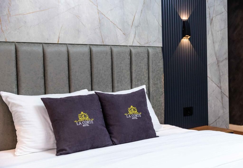 A hotel room with a bed, two pillows with a logo, a headboard, and a wall-mounted light fixture.
