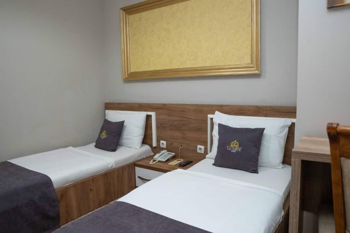 Une chambre d’hôtel avec deux lits simples, une tête de lit en bois, une table de chevet avec un téléphone et une télécommande, et un tableau encadré au mur.