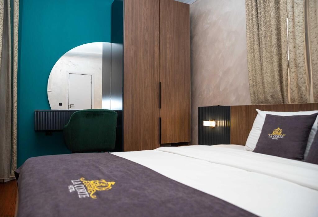 A hotel room with a bed, a green chair, a wooden wardrobe, a mirror, and a wall with curtains.
