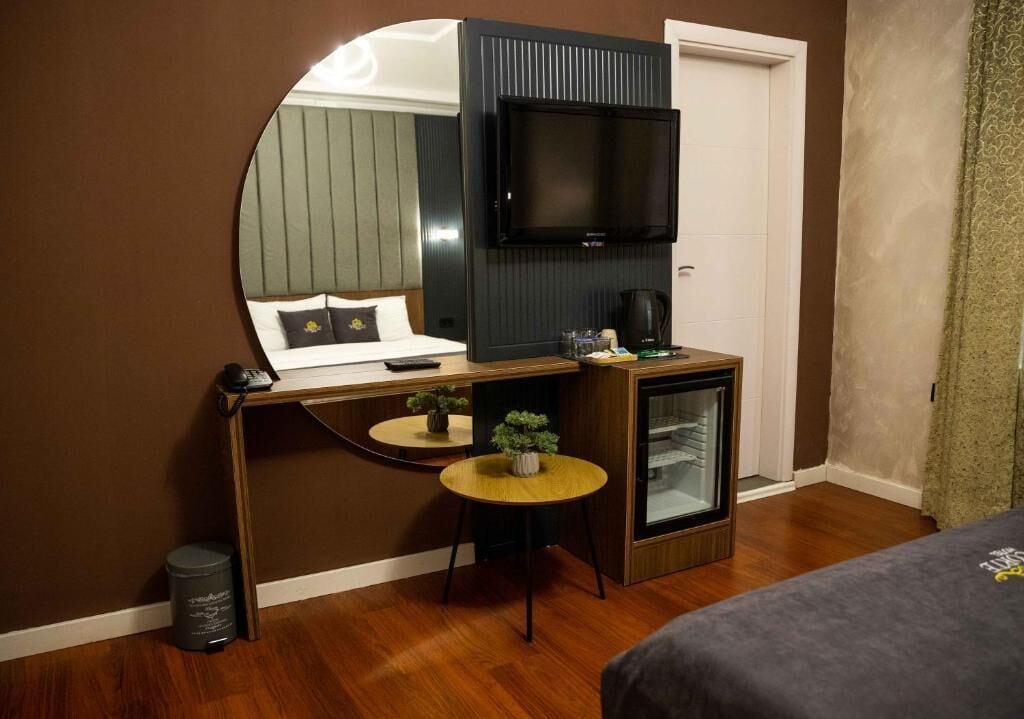 A compact hotel room with a bed, a desk with a computer, a television, a small refrigerator, and a round table with a plant.