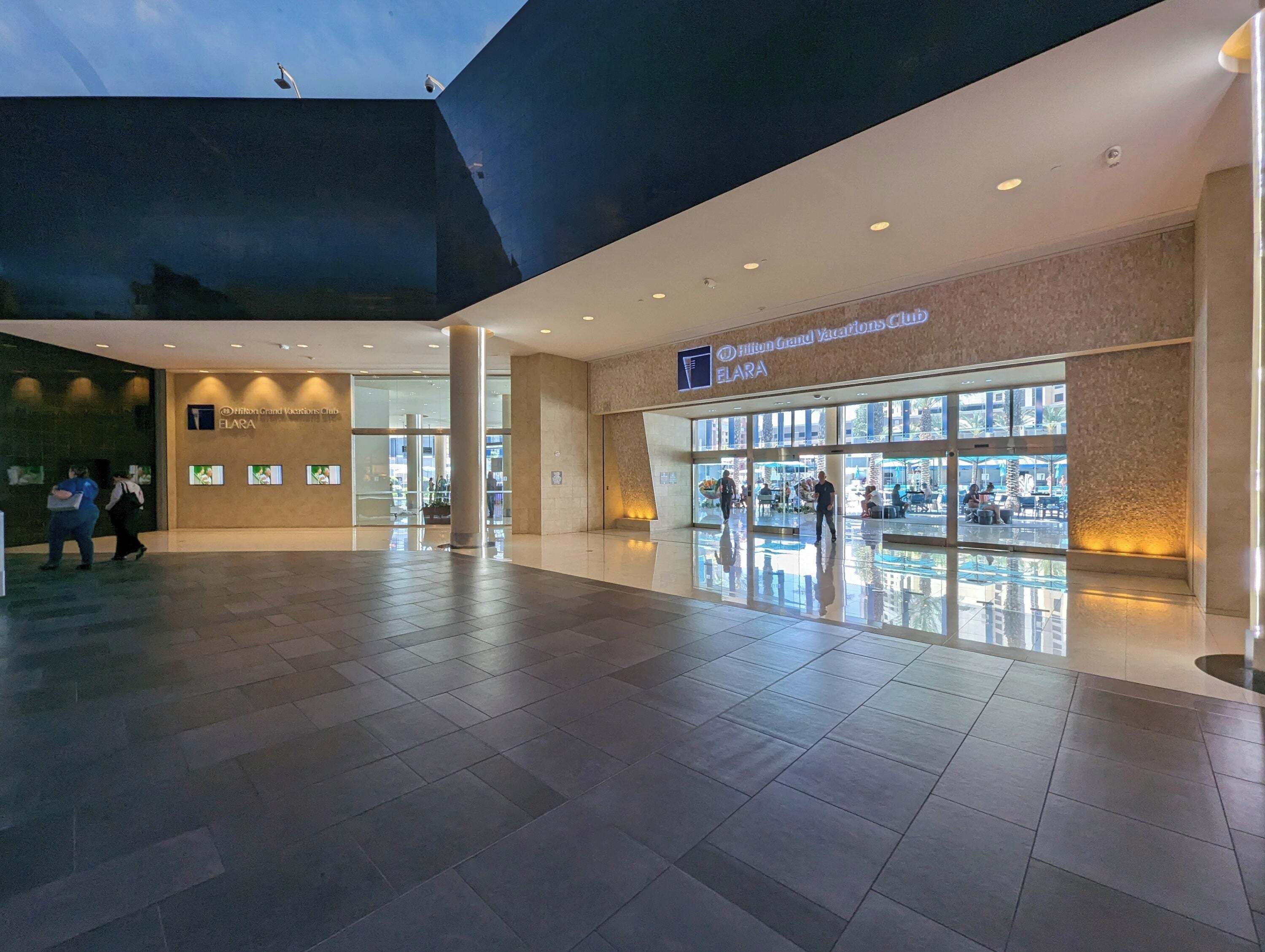 A spacious lobby with a modern design, a visible entrance to the Hilton Grand Vacations Club ELARA, and a large glass window offering a view of the outdoor area.