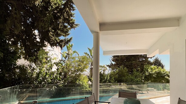 A poolside area with a glass railing, a table with chairs, and a view of a swimming pool. - Carthage Villa 3BR Luxury Pool Garden (Carthage)