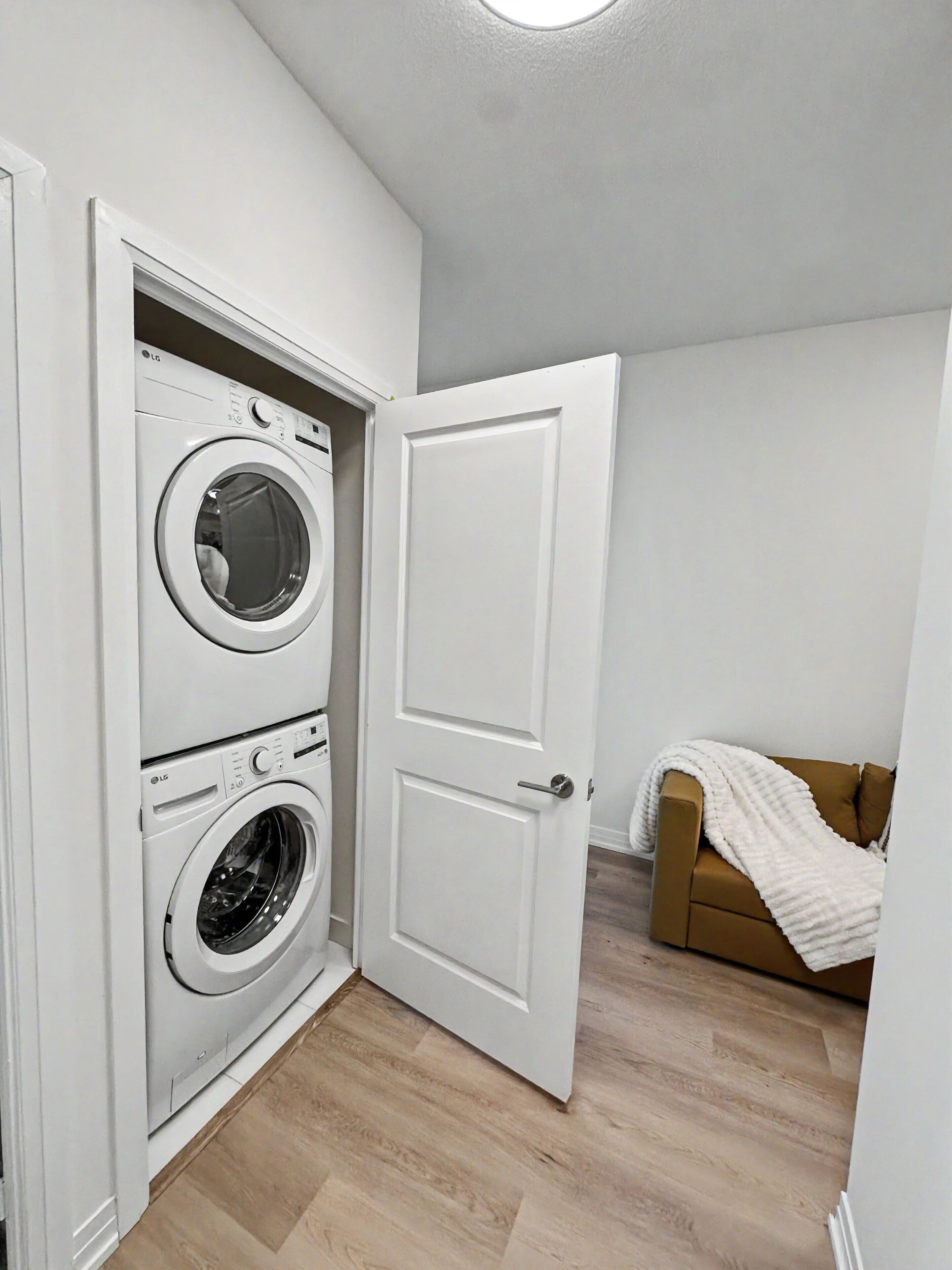 A laundry room with two stacked washing machines, a white door, and a brown couch with a white blanket.