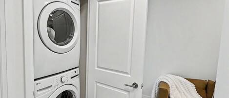 A laundry room with two stacked washing machines, a white door, and a brown couch with a white blanket.