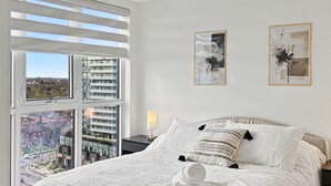 A bedroom with a large bed, a window with blinds, and two framed artworks on the wall. - Modern 2BR+Den Condo near Square One | Sleeps 6 | Mins to Pearson YYZ (Mississauga)