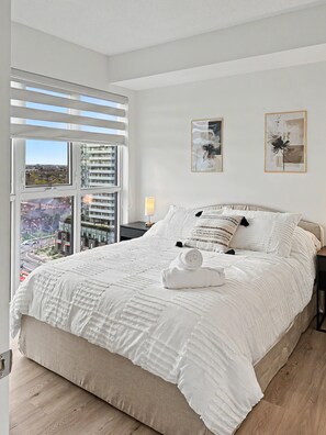 A bedroom with a large bed, a window with blinds, and two framed artworks on the wall.