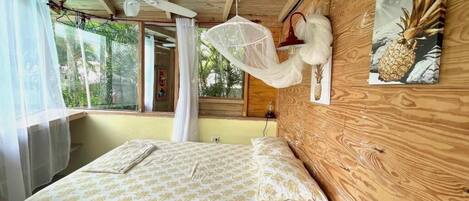 A wooden room with a bed, a ceiling fan, a mosquito net, and a pineapple painting on the wall.