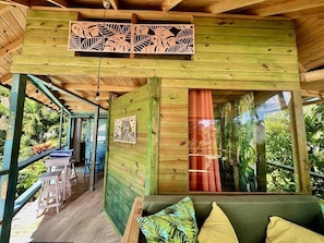 A wooden cabin with a porch, a sofa with cushions, and a large window with a curtain.