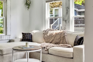 A living room with a sofa, a round table, and a window with a view of a house and a tree.