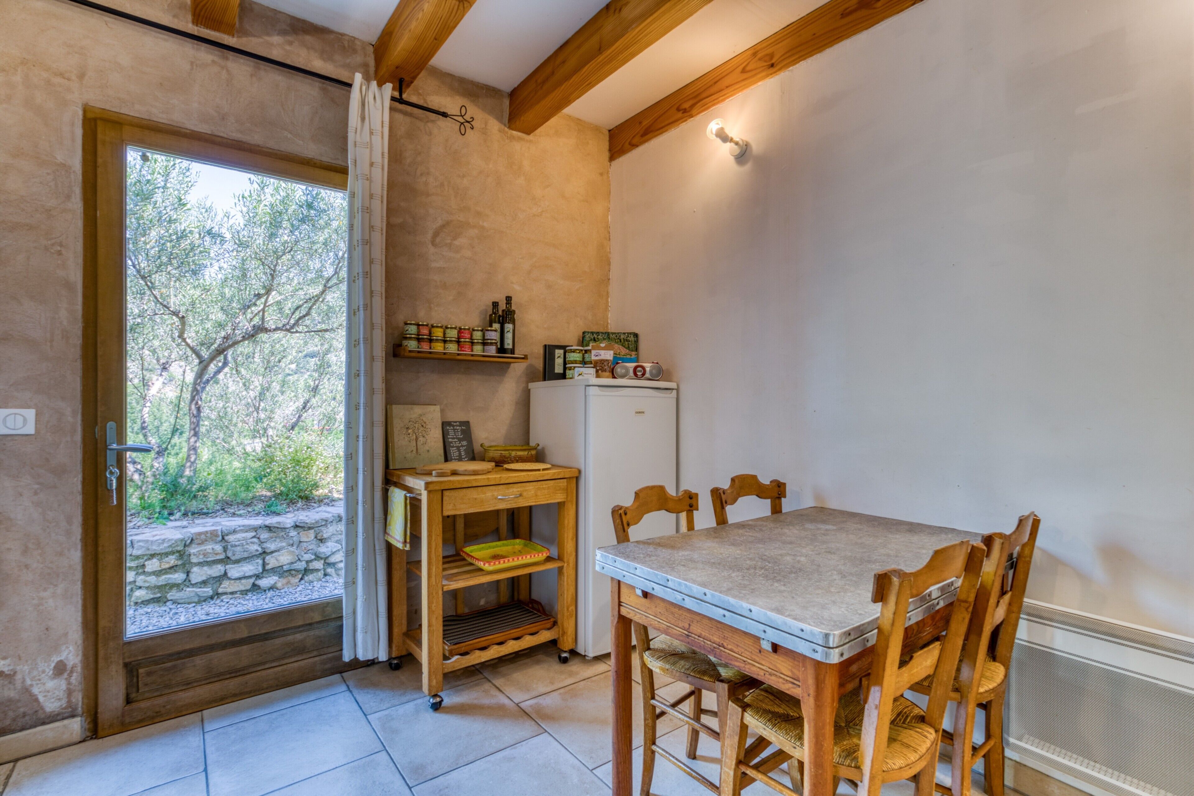 A room with a wooden table, chairs, a small refrigerator, a shelf with bottles, and a window with curtains.