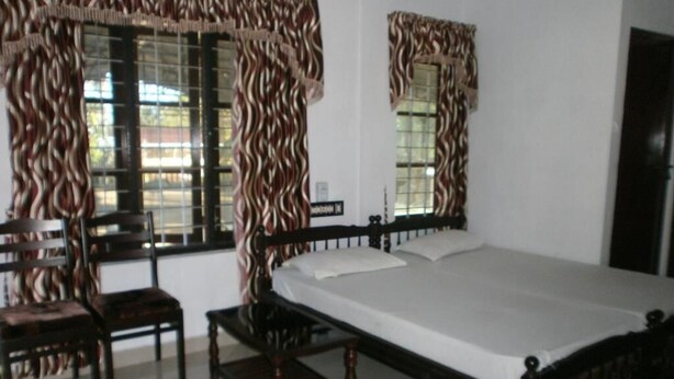 Un dormitorio con cama, dos sillones, una mesita y dos ventanas con cortinas.