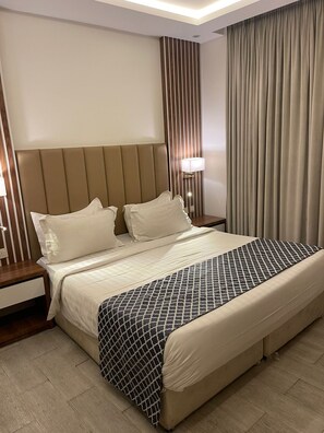 Quarto de hotel com uma cama grande, uma mesa de cabeceira, um abajur e uma cortina.