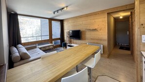 Un interior moderno con una mesa de comedor de madera, sillas blancas, un sofá, un televisor y un amplio ventanal.