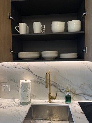 A kitchen with a marble countertop, a gold faucet, and a shelf with white cups and plates. - Stylish Residences Wynwood (Miami)