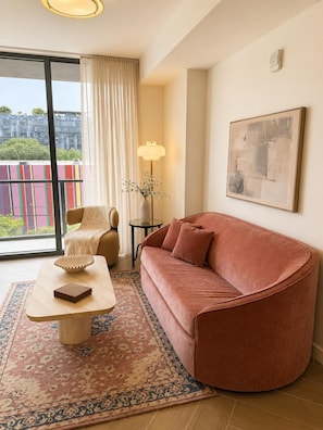 A modern living room with a curved sofa, a coffee table, a floor lamp, and a large window with a view of buildings. - Stylish Residences Wynwood (Miami)