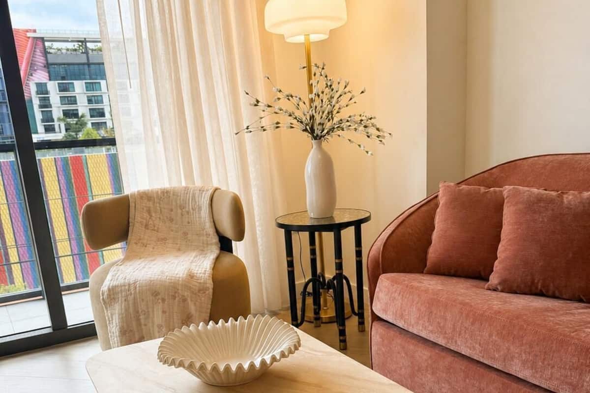 Un salon avec un canapé beige, un fauteuil marron, une table basse ornée d’un vase décoratif et d’un livre, un lampadaire et une vue sur les immeubles à travers une grande fenêtre.
