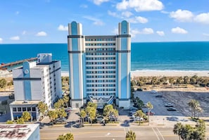 A high-rise hotel with a modern design, a parking lot, and a beach view. - Direct Oceanfront Condo with Stunning Views! (Myrtle Beach)