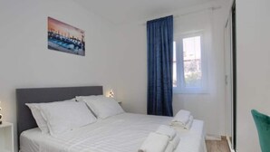 A bedroom with a bed, white bedding, a nightstand, a wall-mounted picture, a window with blue curtains, and a green chair. - Two Bedroom Apartment With Terrace (Pula)