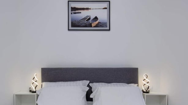 A bed with white linens, two white pillows, a dark headboard, and a framed picture of a lake with a boat on the wall. - Two Bedroom Apartment With Terrace (Pula)