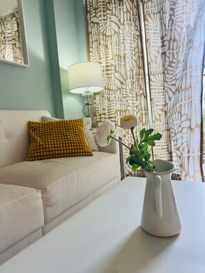 Una sala de estar con un sofá blanco, un cojín amarillo estampado, un jarrón blanco con una flor y una cortina con diseño.
