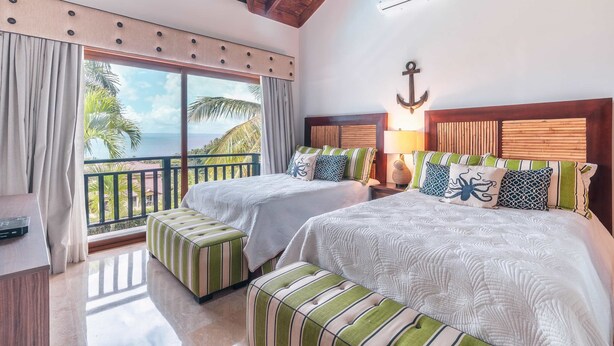Habitación de hotel con dos camas, un balcón con vista a palmeras y una decoración de ancla en la pared.