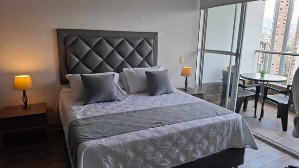 Een hotelkamer met een groot bed, twee bedlampen, een balkon met een tafel en stoelen, en uitzicht over de stad.