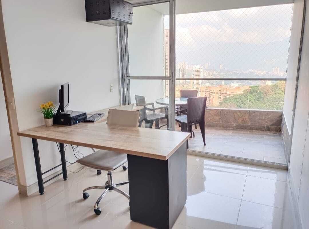 Un espacio de oficina moderno con escritorio, silla y computadora, una planta pequeña y vistas de la ciudad a través de grandes ventanales.