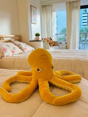 Ein Hotelzimmer mit Bett, einem Fenster mit Vorhängen, einem kleinen Tisch mit Pflanze und einem farbenfrohen Kissen.