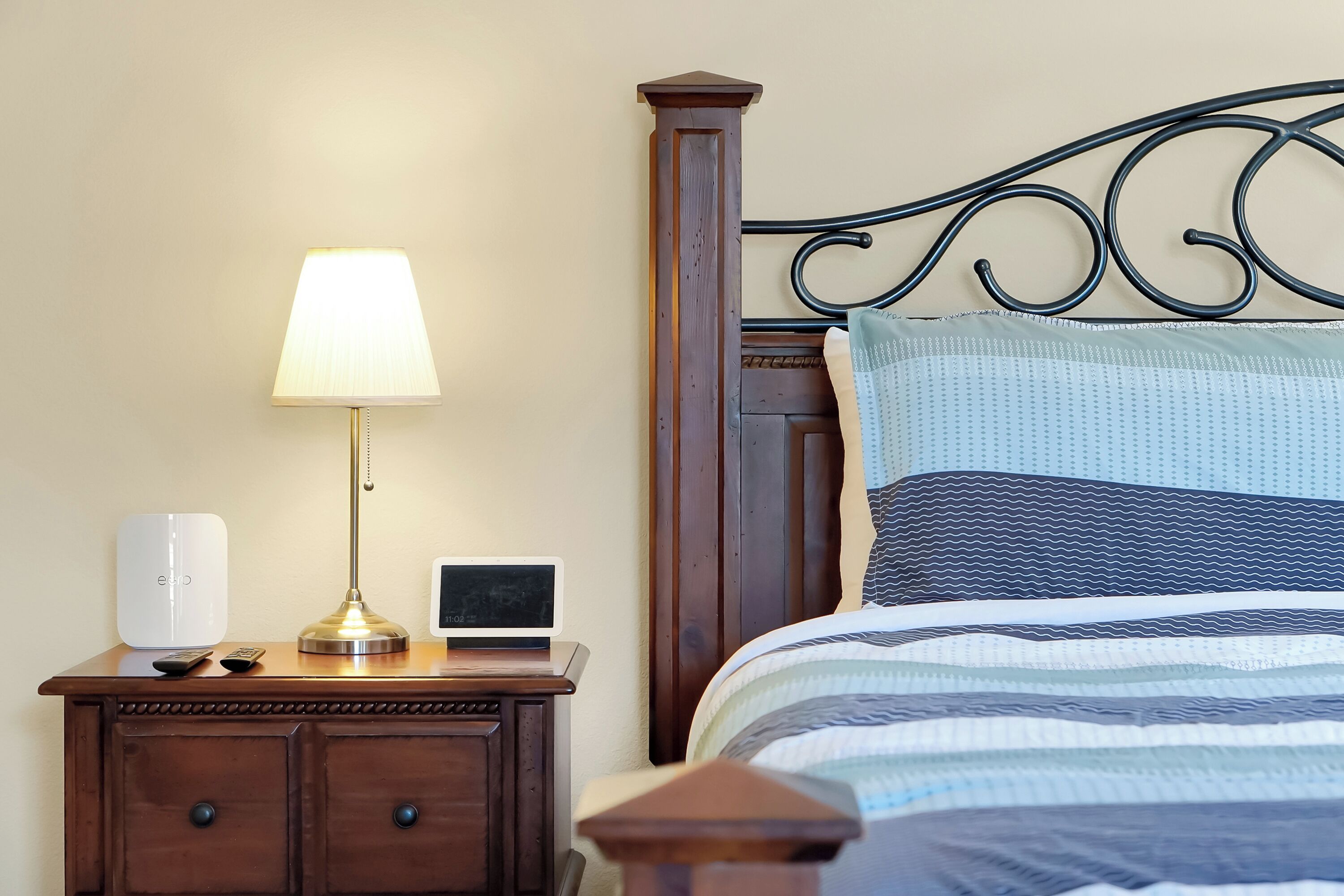A neatly made bed with a striped bedspread, a bedside lamp, a small table with a lamp, a speaker, and a remote control.