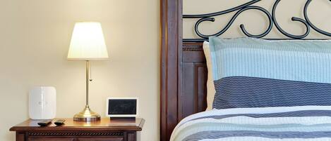 A neatly made bed with a striped bedspread, a bedside lamp, a small table with a lamp, a speaker, and a remote control.