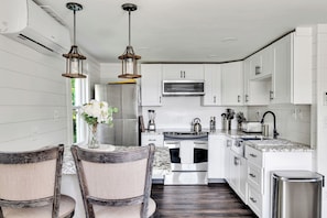 A modern kitchen with white cabinets, stainless steel appliances, and a marble countertop. There are two wooden chairs and a vase with flowers on the table.