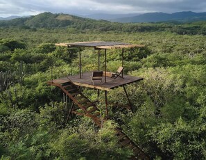 A wooden deck with a chair and a table, surrounded by dense vegetation and hills in the background.