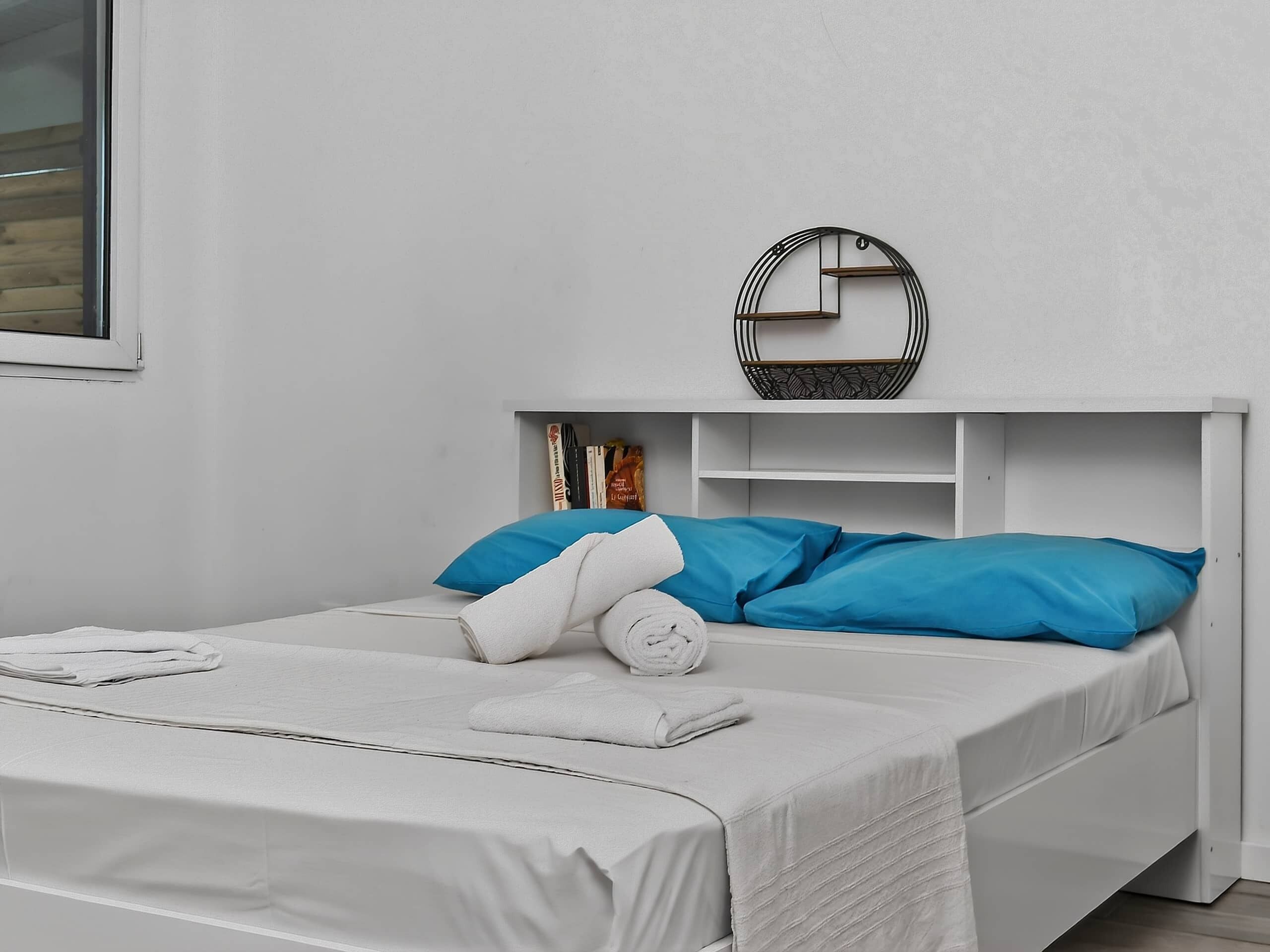 A single-bed room with a white headboard, a bedside table, a wall-mounted shelf with books, and a window.