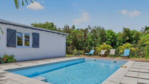 En swimmingpool med solstolar, en liten byggnad med fönsterluckor och frodig grönska.