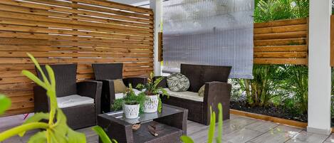 A modern outdoor seating area with wicker furniture, a glass coffee table, and a view of greenery through a window.
