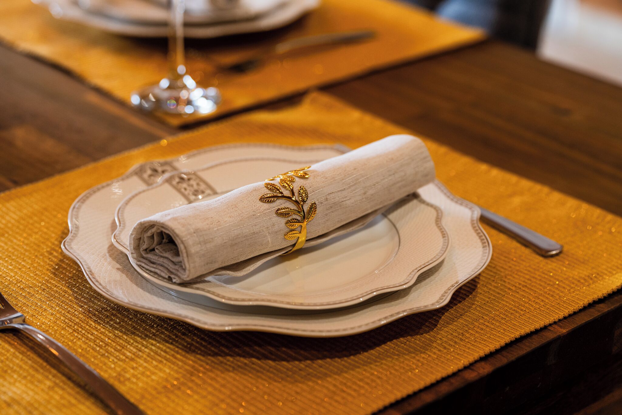 Une serviette pliée avec soin, ornée d’un motif doré, est posée sur une assiette, à côté d’une fourchette et d’un verre, sur une table en bois recouverte d’une nappe texturée.
