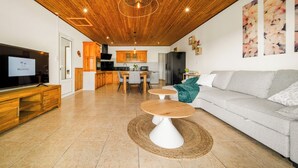 A modern living room with a wooden ceiling, a grey sofa, a TV, and a kitchen in the background.