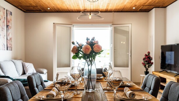 A dining area with a wooden table set for six, a vase with flowers, and a television in the background.