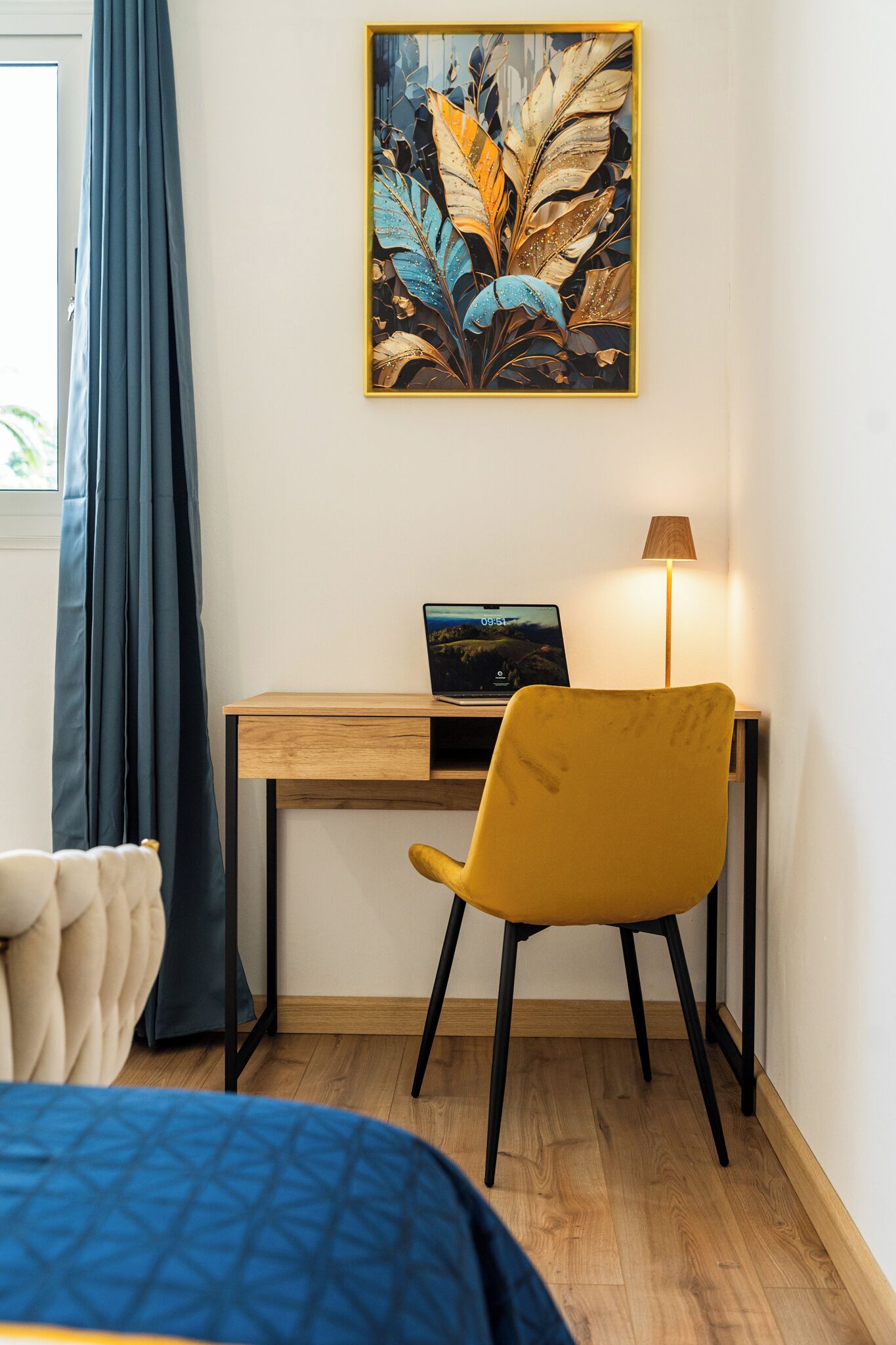 A room with a bed, a desk with a laptop, a chair, a lamp, and a painting on the wall.