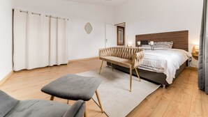 A bedroom with a bed, a chair, a small table, a mirror, and a window with curtains.