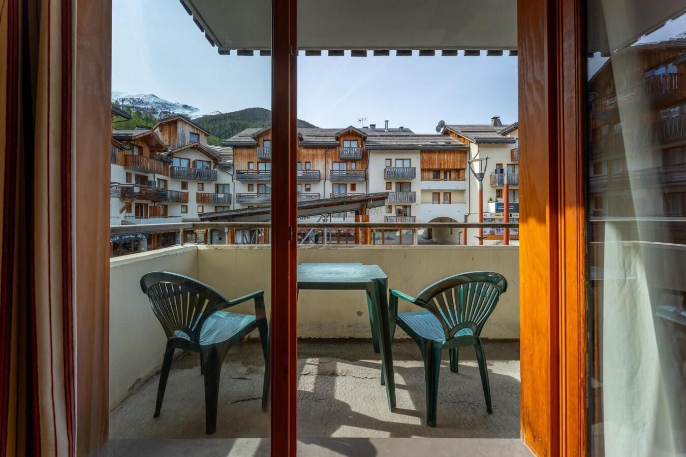 A balcony with two chairs and a table, overlooking a row of wooden buildings with balconies.