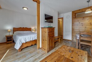 Ein Bett mit Holzgestell und Kopfteil, ein Holzschrank, ein Holztisch, eine Bank und ein an der Wand befestigtes Fernsehgerät.