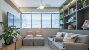 Ein modernes Wohnzimmer mit Sofa, Hocker, Bücherregal und einem großen Fenster.