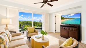 Un salón con televisor, sofá, sillón, mesa de centro y vistas a un paisaje tropical a través de amplios ventanales.