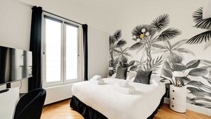 A hotel room with a bed, a desk, a chair, a TV, a window with curtains, and a wall with a tropical leaf mural. - Spacious Appartement 2br/7p - Nogent-sur-marne (Nogent-sur-Marne)