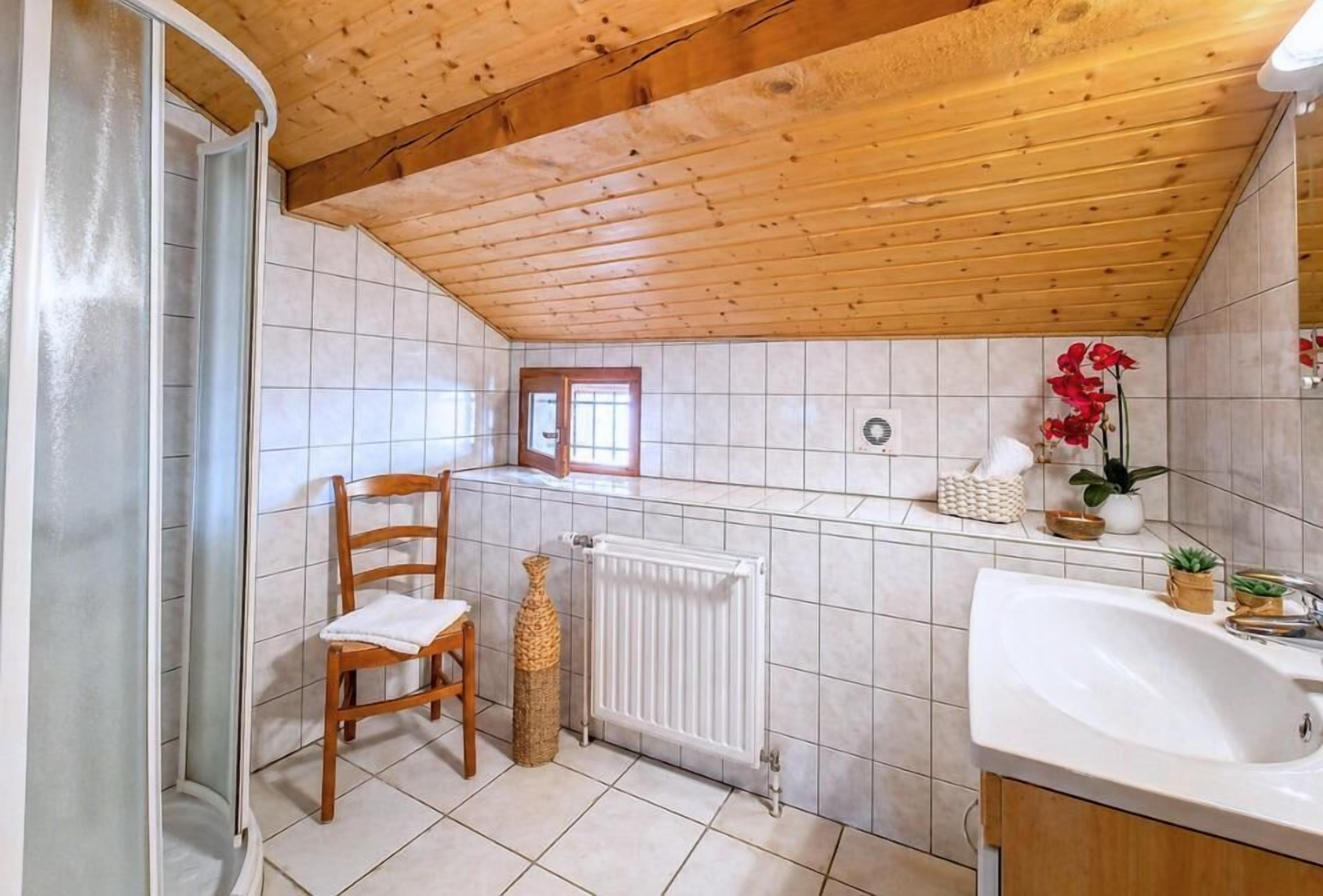 Une salle de bain avec un plafond en bois, un lavabo blanc, un radiateur, une chaise et une fenêtre.